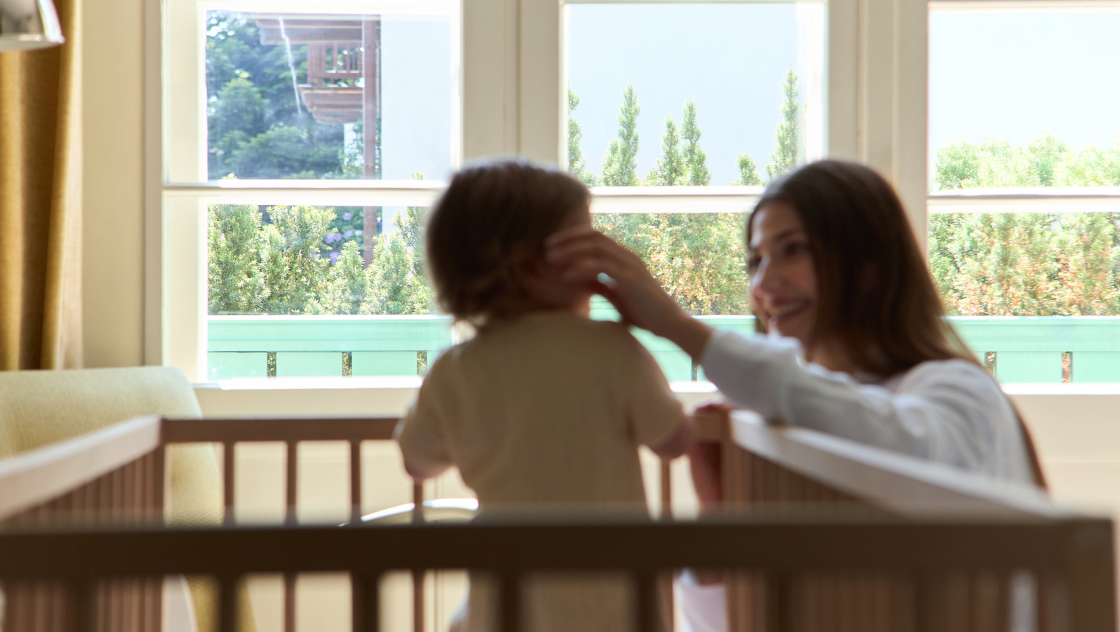 Jasny rodzinny pokój hotelowy z drewnianym łóżeczkiem przy oknie, uśmiechnięta mama przy maluszku, widok na zieleń