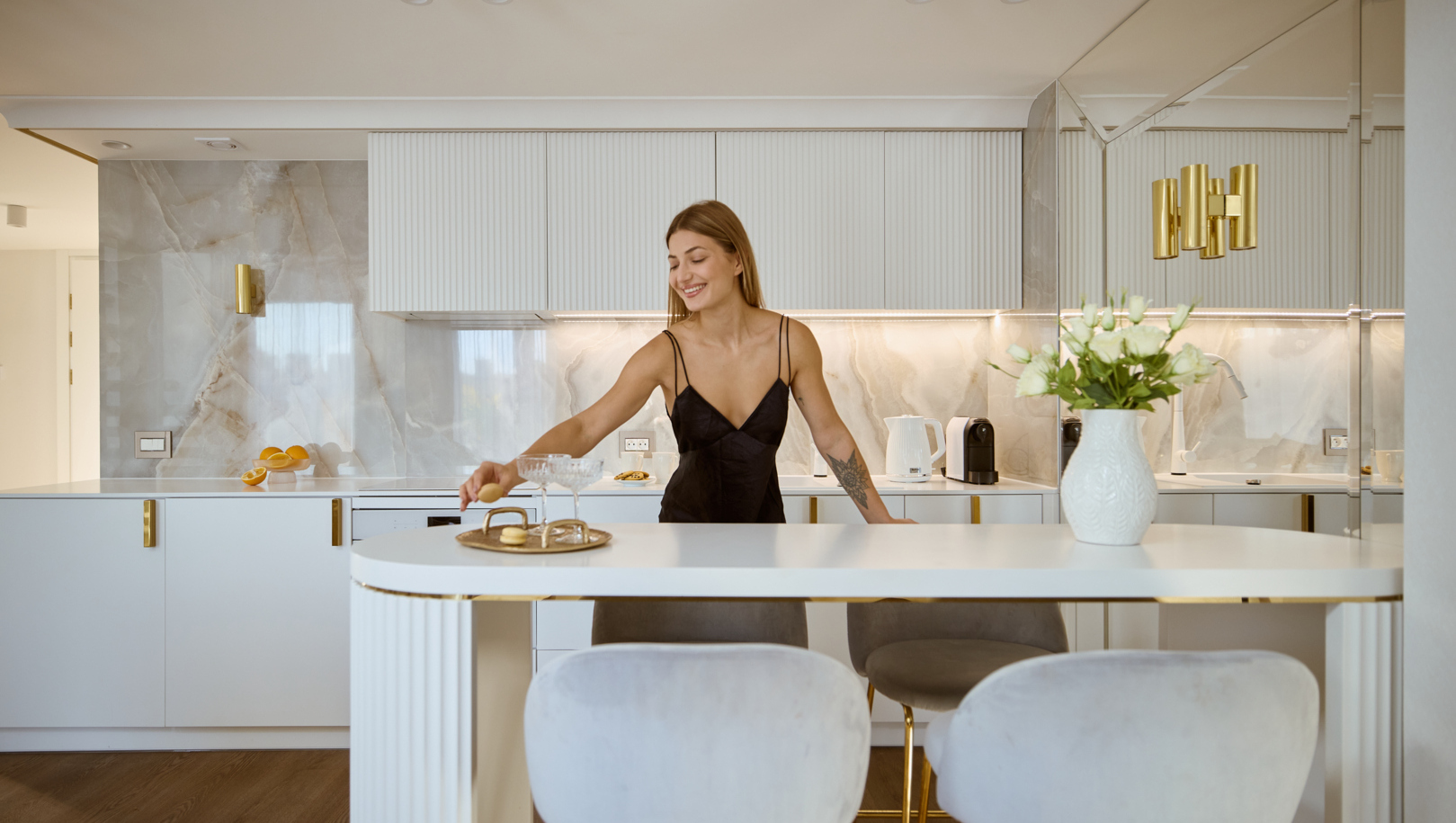 Nowoczesna kuchnia apartamentu z marmurowym panelem, złotymi akcentami, miękkimi hokerami, ekspresem do kawy i kobietą z makaronikiem.