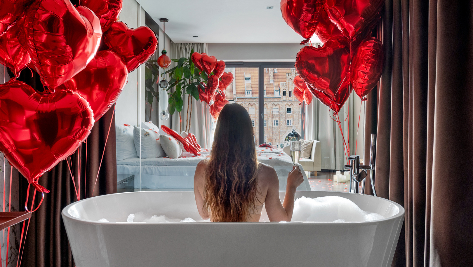 Kobieta w wolnostojącej wannie z pianą, z kieliszkiem szampana; romantyczny apartament z balonami w kształcie serc i widokiem na miasto