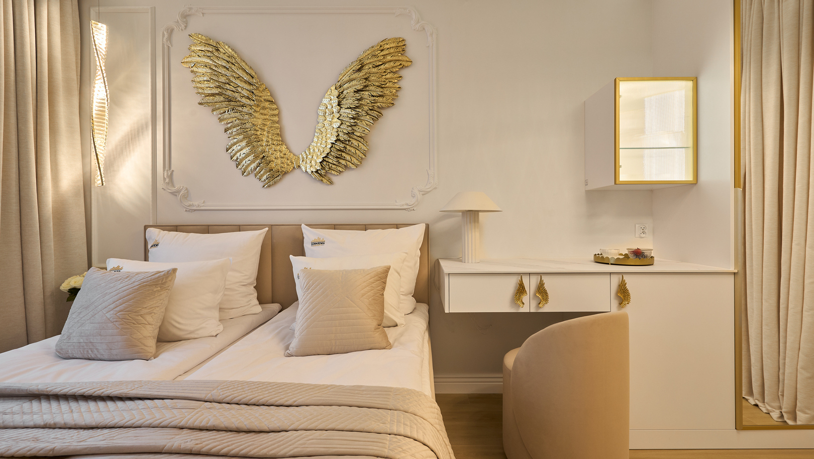 Pokój butikowy z dwoma łóżkami, beżową pikowaną pościelą, złotą rzeźbą skrzydeł, subtelne oświetlenie i biurko ze złotymi akcentami.