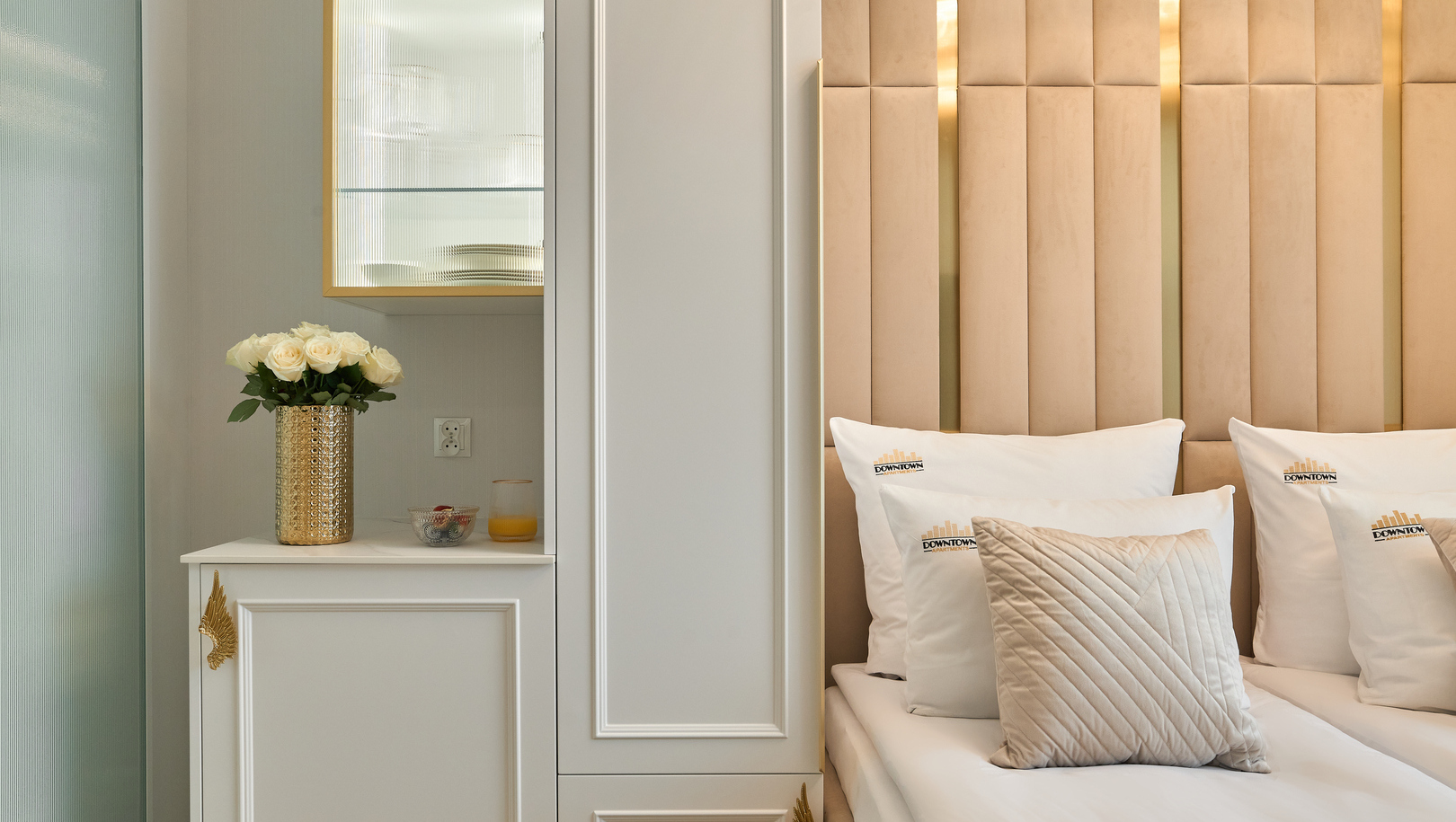 Miękkie łóżka bliźniacze z wysokim beżowym tapicerowanym zagłówkiem, białą pościelą z poduszkami z logo, pikowaną poduszką i złotym wazonem z róż.