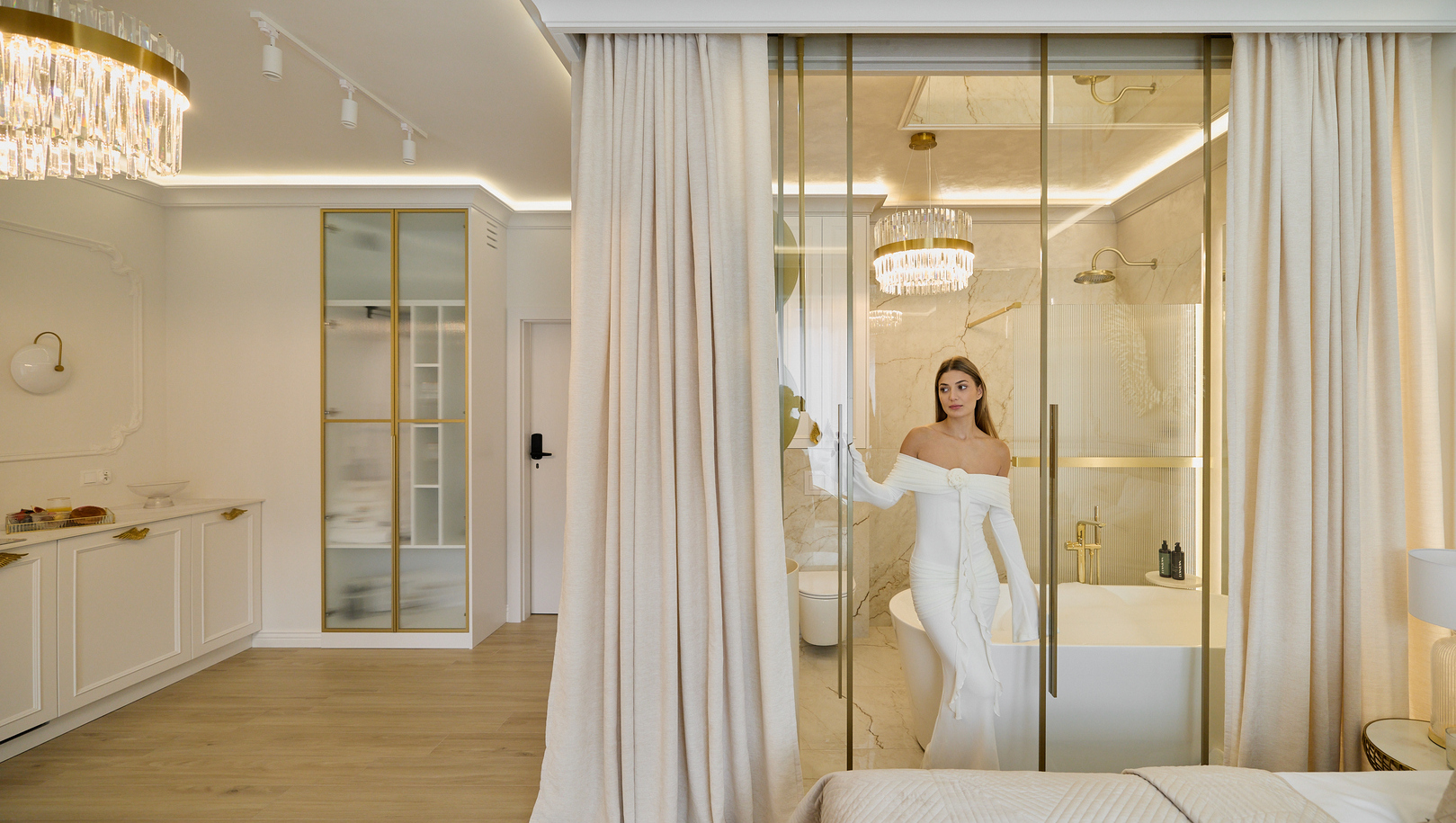 Luksusowy apartament z przeszkloną marmurową łazienką, wolnostojącą wanną, złotymi armaturami i żyrandolem; kobieta w białej sukni przy drzwiach.