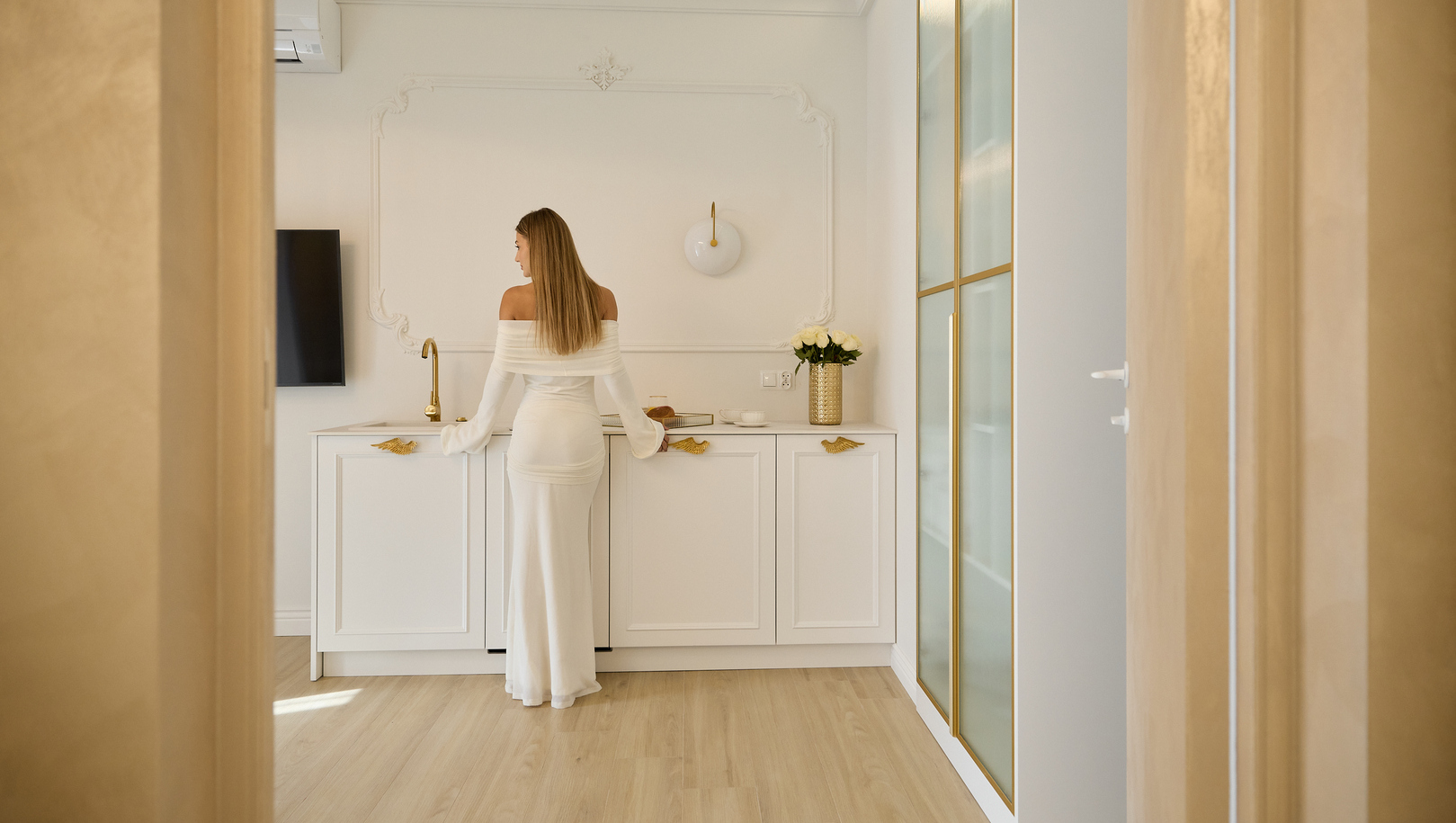 Kobieta w eleganckiej białej sukni przy aneksie w butikowym apartamencie, złote baterie, szafa z matowym szkłem i wazon z białymi różami