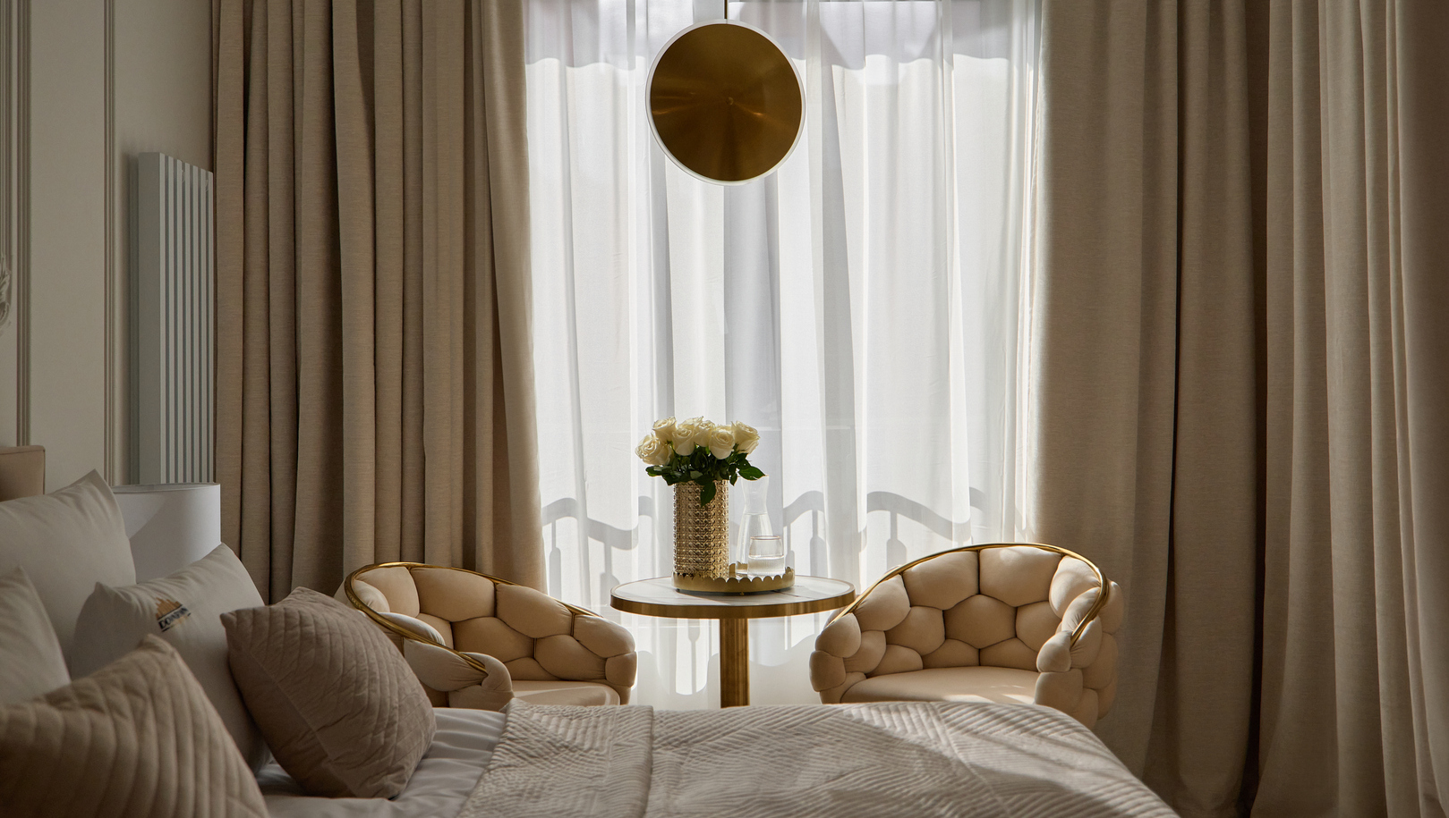 Pokój butikowego hotelu w miękkim świetle, łóżko z pikowanymi poduszkami, dwa beżowe pikowane fotele i okrągły stolik z białymi różami.