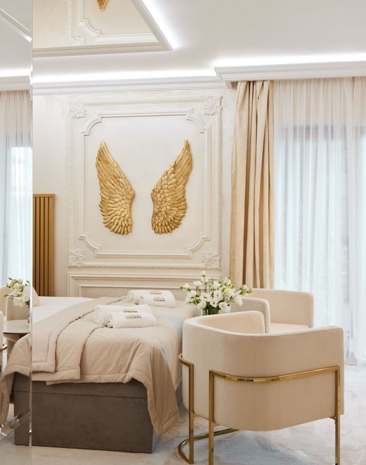Luksusowy pokój hotelowy z miękkim łóżkiem, złotą rzeźbą skrzydeł na ścianie, kremowymi fotelami i delikatnym światłem.