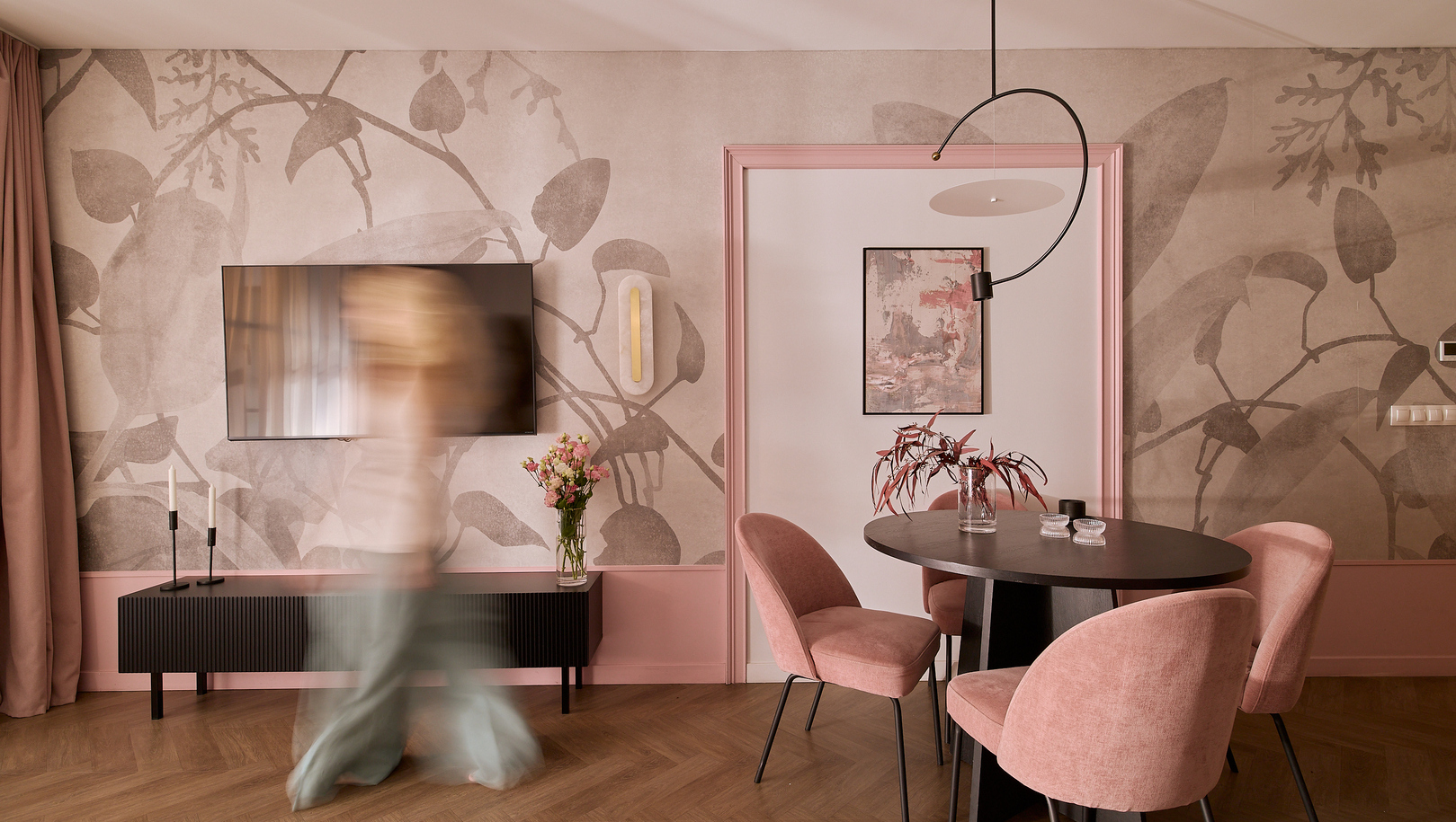 Salon w butikowym apartamencie z kwiatową tapetą, telewizorem, okrągłym ciemnym stołem, różowymi aksamitnymi krzesłami i wazonem; gość przechodzi.