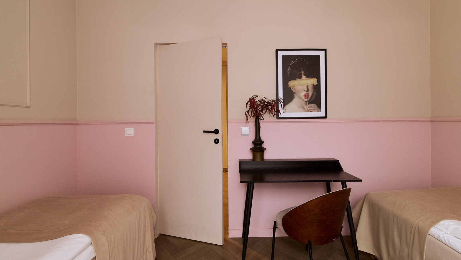 Pokój z dwoma łóżkami jednoosobowymi, pastelowe różowe ściany, biurko, krzesło, obraz i wazon z rośliną.