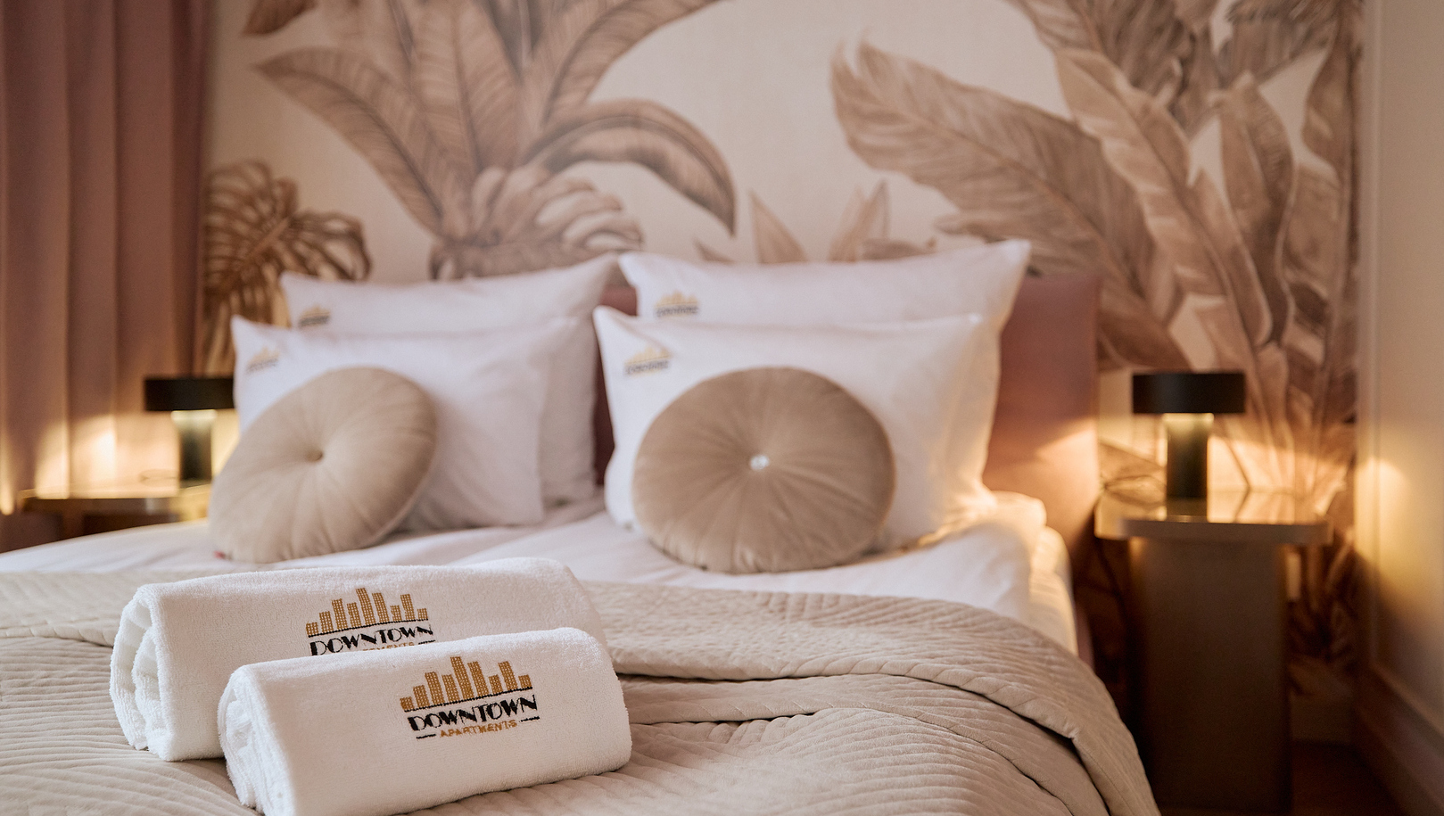 Łóżko dwuosobowe z pikowanym beżowym narzutem, okrągłymi poduszkami, zwiniętymi ręcznikami z logo i ciepłym oświetleniem przy botanicznej tapecie.