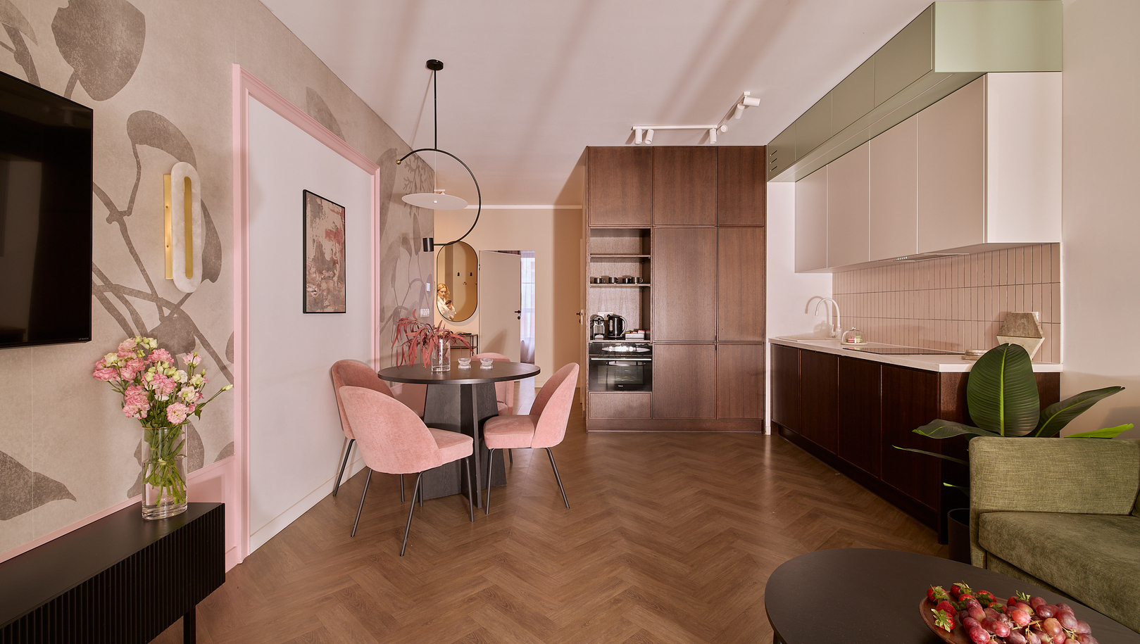 Aneks apartamentu z jadalnią: okrągły stół, różowe aksamitne krzesła, jodełkowa podłoga, sofa, telewizor i nowoczesne AGD.