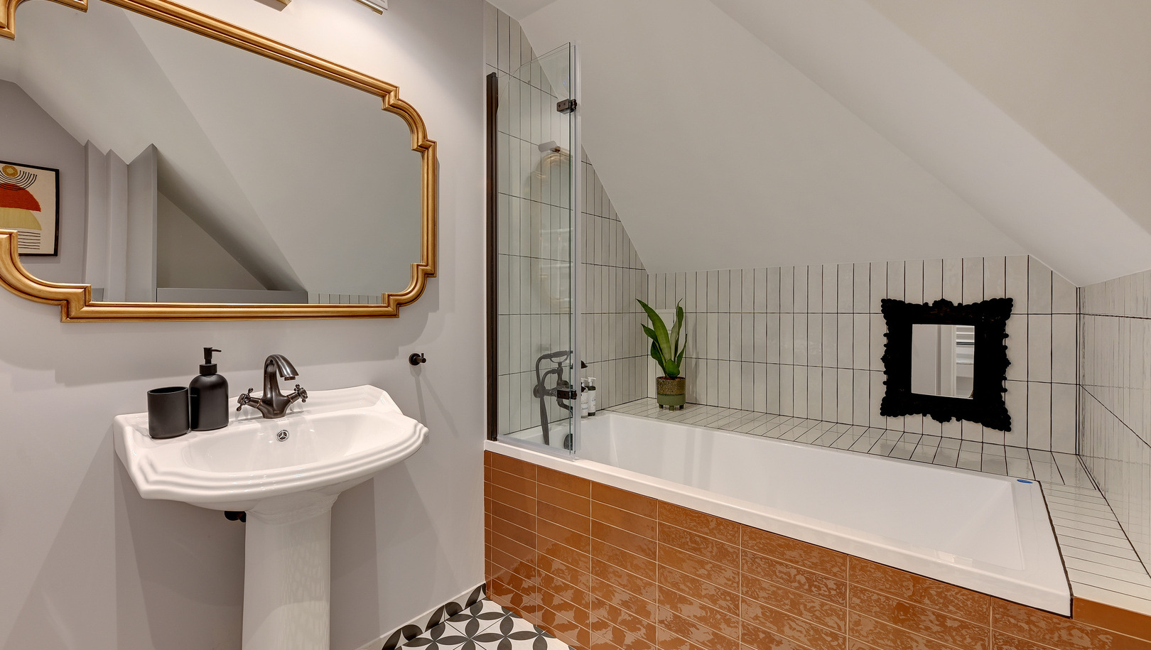 Łazienka na poddaszu z wanną i szklaną ścianką prysznicową, front wanny w pomarańczowych płytkach, umywalka stojąca, złota rama lustra, wzorzysta posadzka.