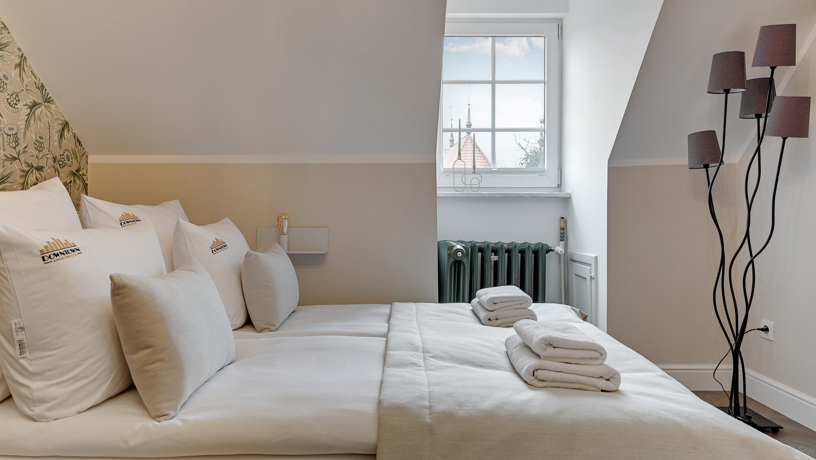 Przytulny pokój na poddaszu ze skosem sufitu, białą pościelą, złożonymi ręcznikami, kaloryferem i oknem z widokiem na miasto