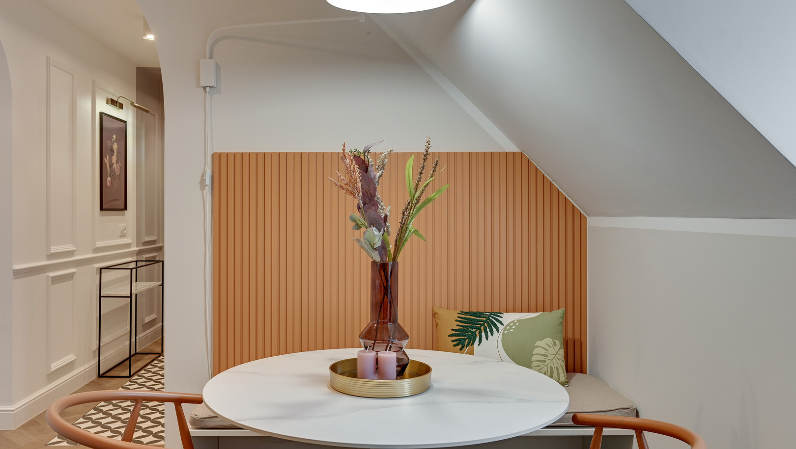 Przytulny kącik jadalniany na poddaszu z okrągłym białym stołem, wbudowaną ławką, pomarańczową boazerią, wazonem z suchymi kwiatami