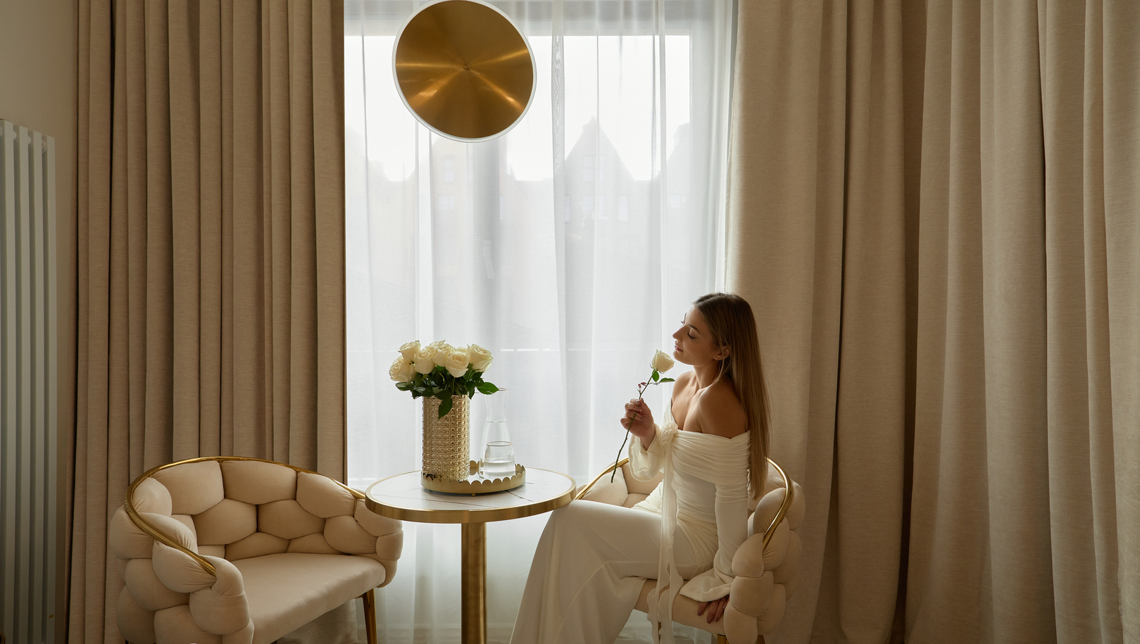 Luksusowy kącik hotelowy z miękkimi kremowymi fotelami, złotym okrągłym stolikiem, wazonem białych róż i gościem wąchającym różę przy lekkich zasłonach.