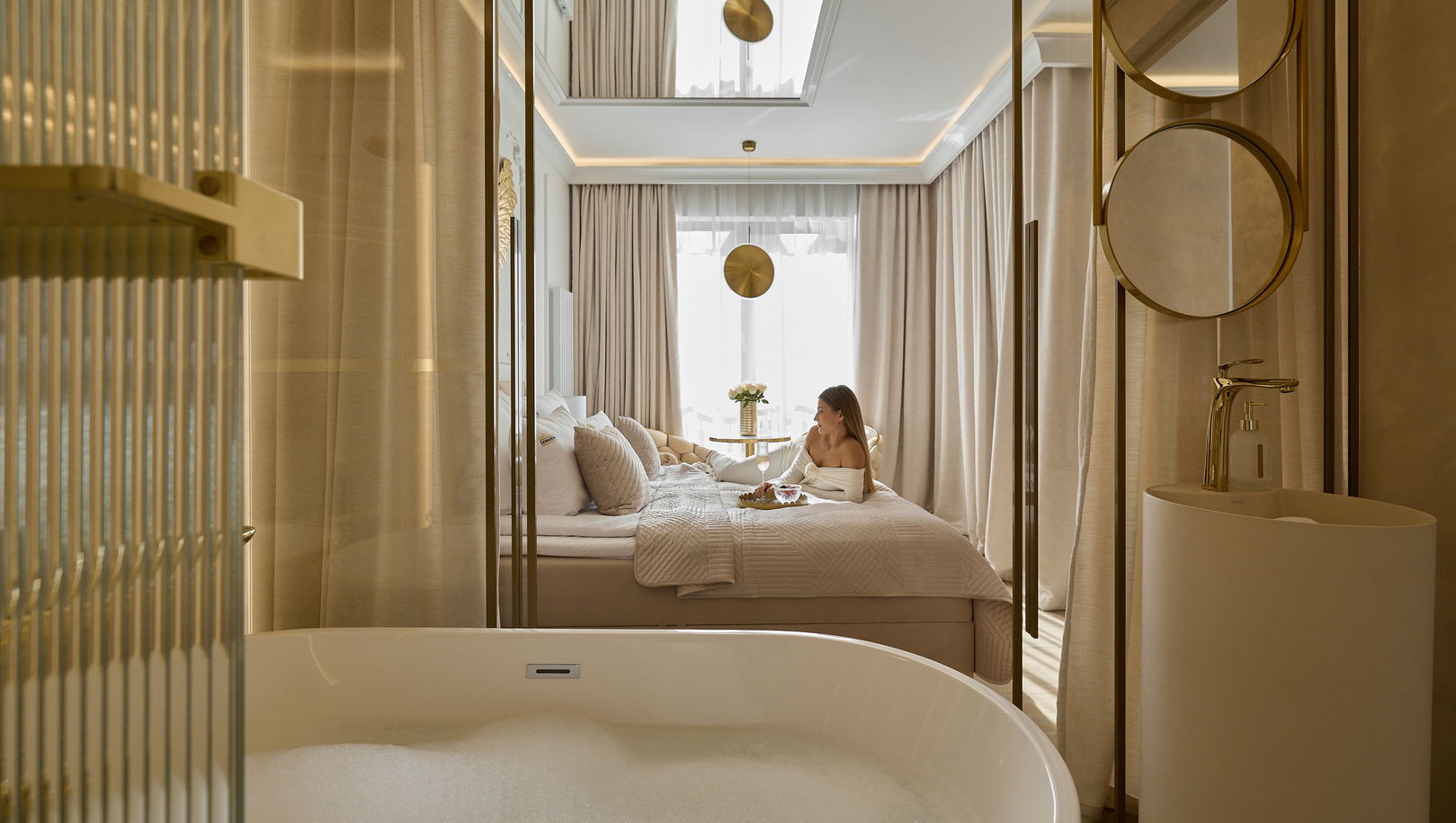 Wanna wolnostojąca z pianą otwarta na beżowy apartament z złotymi akcentami, miękkim łóżkiem, wysokimi zasłonami i gościem przy śniadaniu