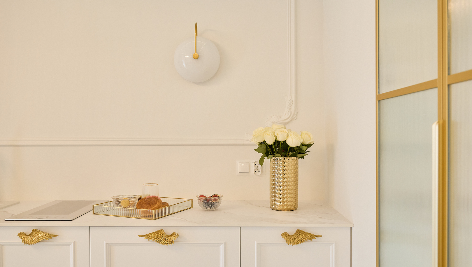 Biały marmurowy blat i szafki ze złotymi akcentami, białe róże w złotym wazonie, taca z ciastkami, szklanka i kinkiet