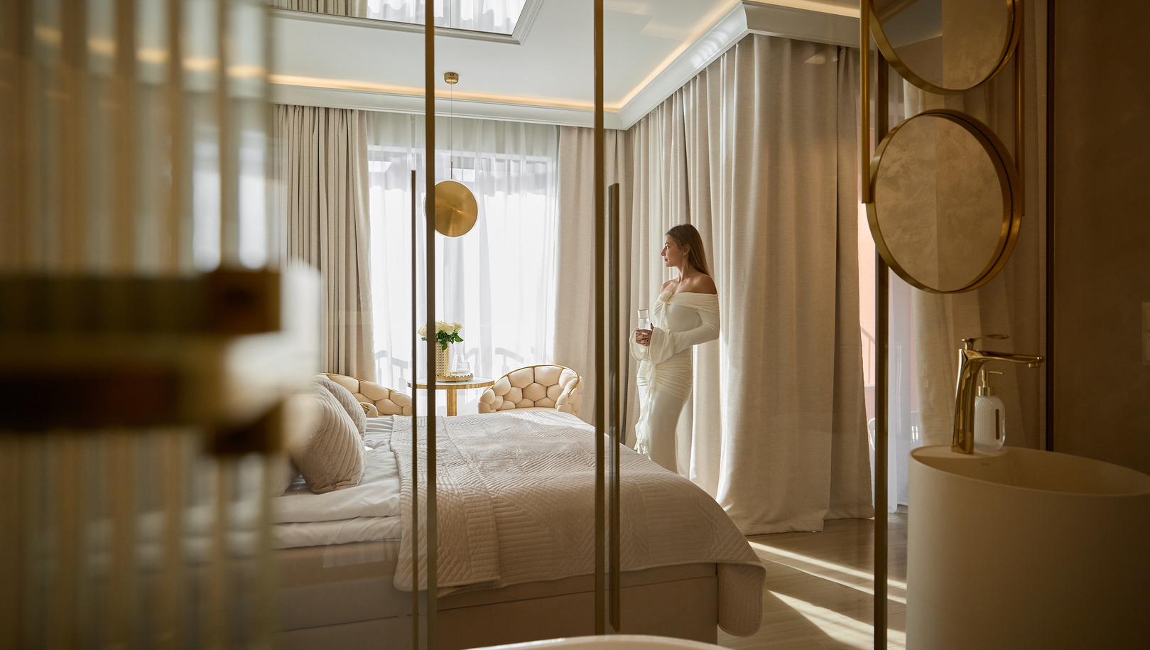Luksusowy apartament z dużym łóżkiem, pikowanymi fotelami i stolikiem z kwiatami, wolnostojąca wanna i złote baterie, kobieta w białej sukni patrzy w światło.
