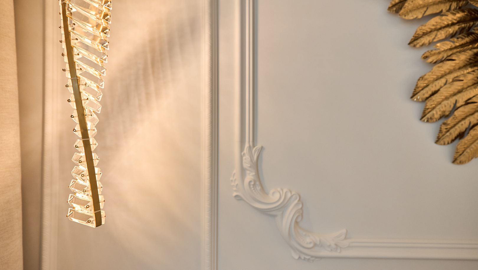 Ciepła kryształowa lampa wisząca rzucająca miękkie światło na ozdobną białą sztukaterię i złotą dekorację listków