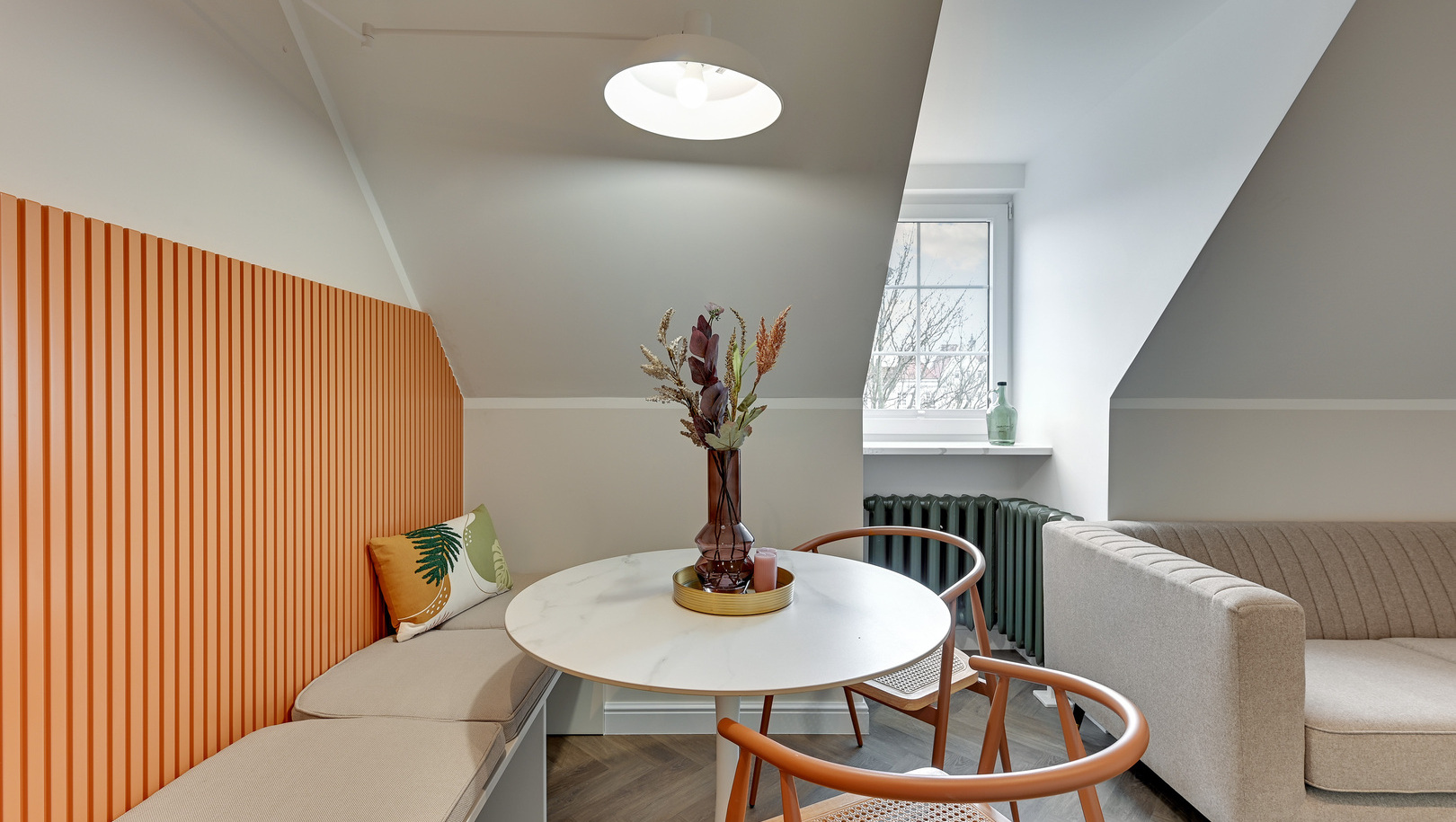 Przytulny kącik jadalny w hotelu pod skośnym sufitem z pomarańczową lamperią, okrągłym marmurowym stołem, rattanowymi krzesłami, wazonem i sofą