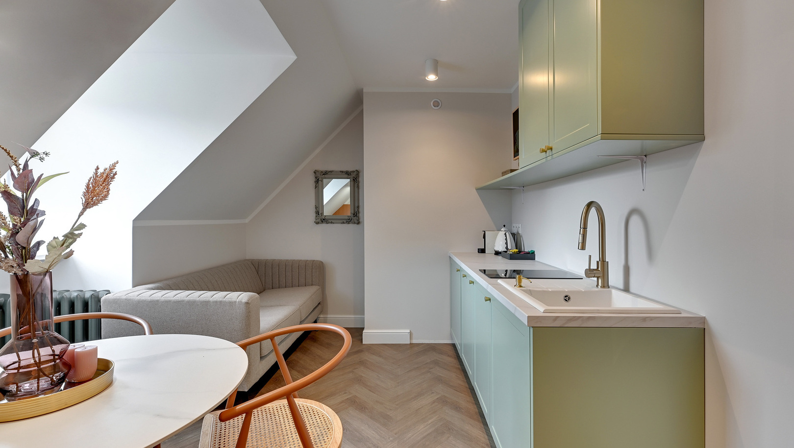 Aneks kuchenny na poddaszu z miętowymi szafkami, mosiężną baterią przy zlewie, okrągły stół z wazonem, rattanowe krzesło, beżowa sofa pod oknem dachowym