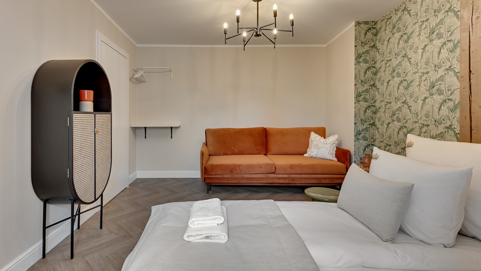 Pokój dwuosobowy z łóżkiem i złożonymi ręcznikami, pomarańczową welurową sofą, zdobną tapetą, nowoczesnym żyrandolem i jodełką.