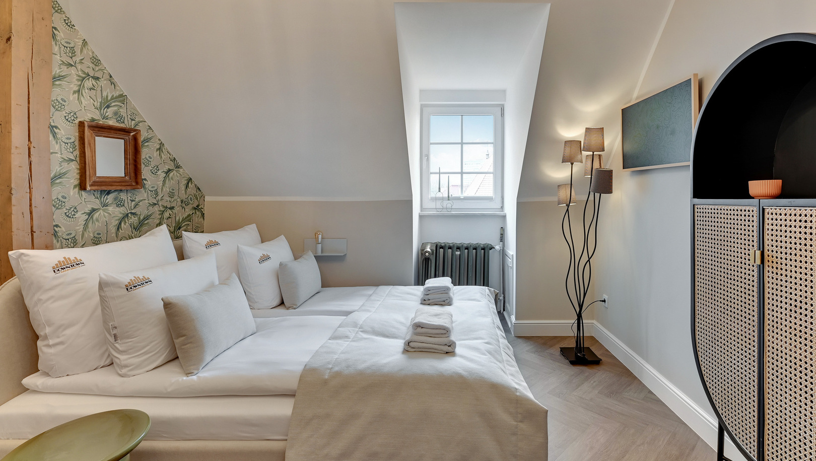 Butikowy pokój na poddaszu z pochyłym sufitem, białą pościelą, złożonymi ręcznikami na łóżku, kwiatową tapetą, oknem i rattanową szafką.