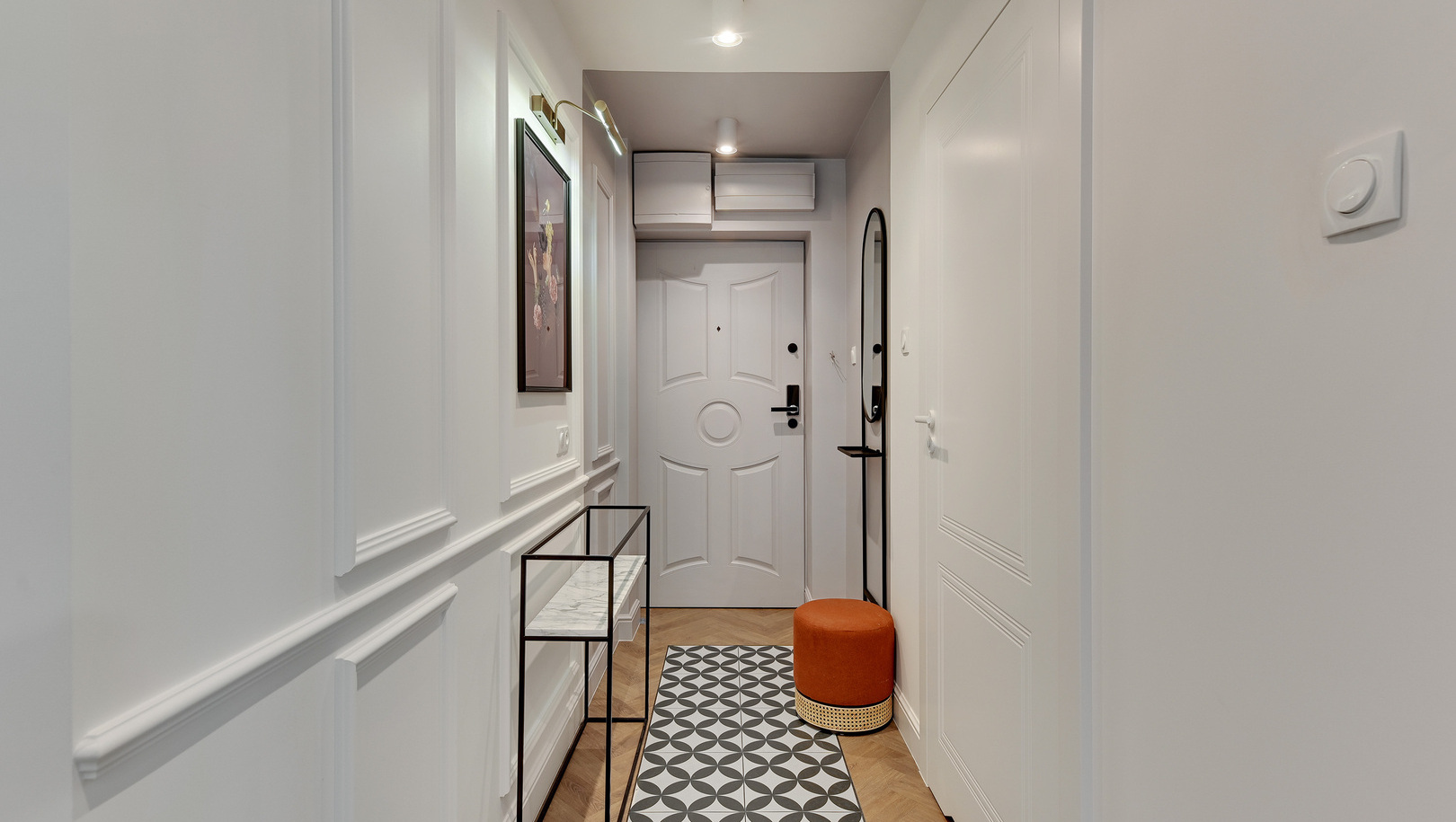 Jasne białe wejście hotelowe z boazerią, geometrycznym dywanikiem, marmurową konsolą, okrągłym lustrem i pomarańczowym pufem.