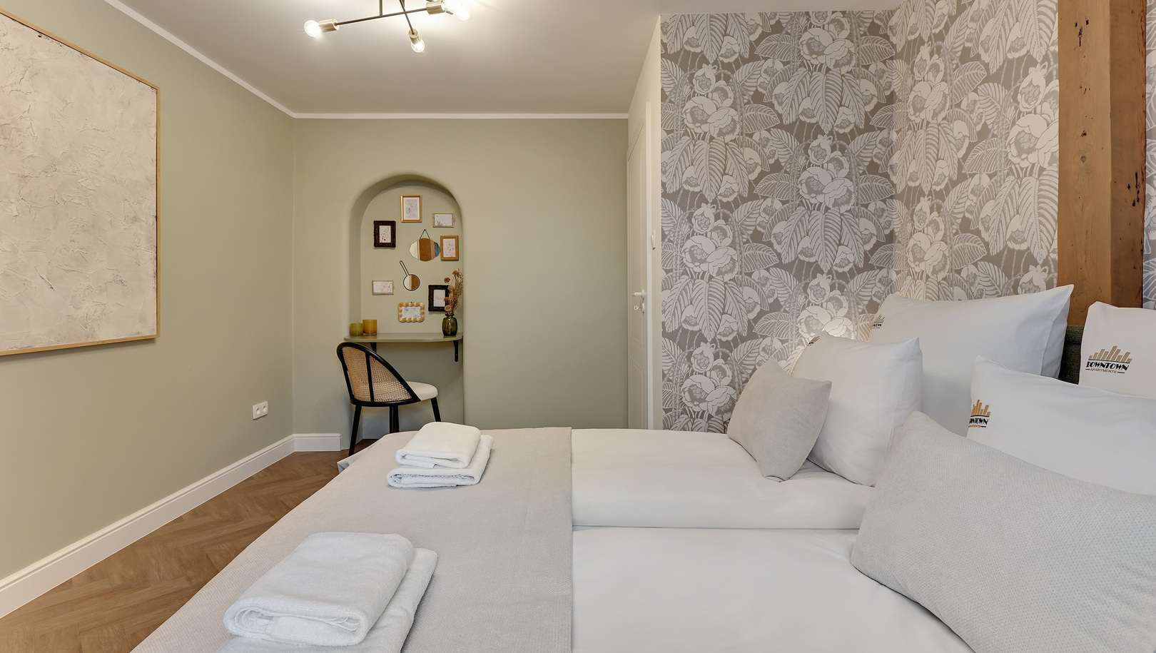 Dwa łóżka z białą pościelą i ręcznikami, dekoracyjna kwiatowa tapeta, drewniana belka i przytulne biurko w wnęce.