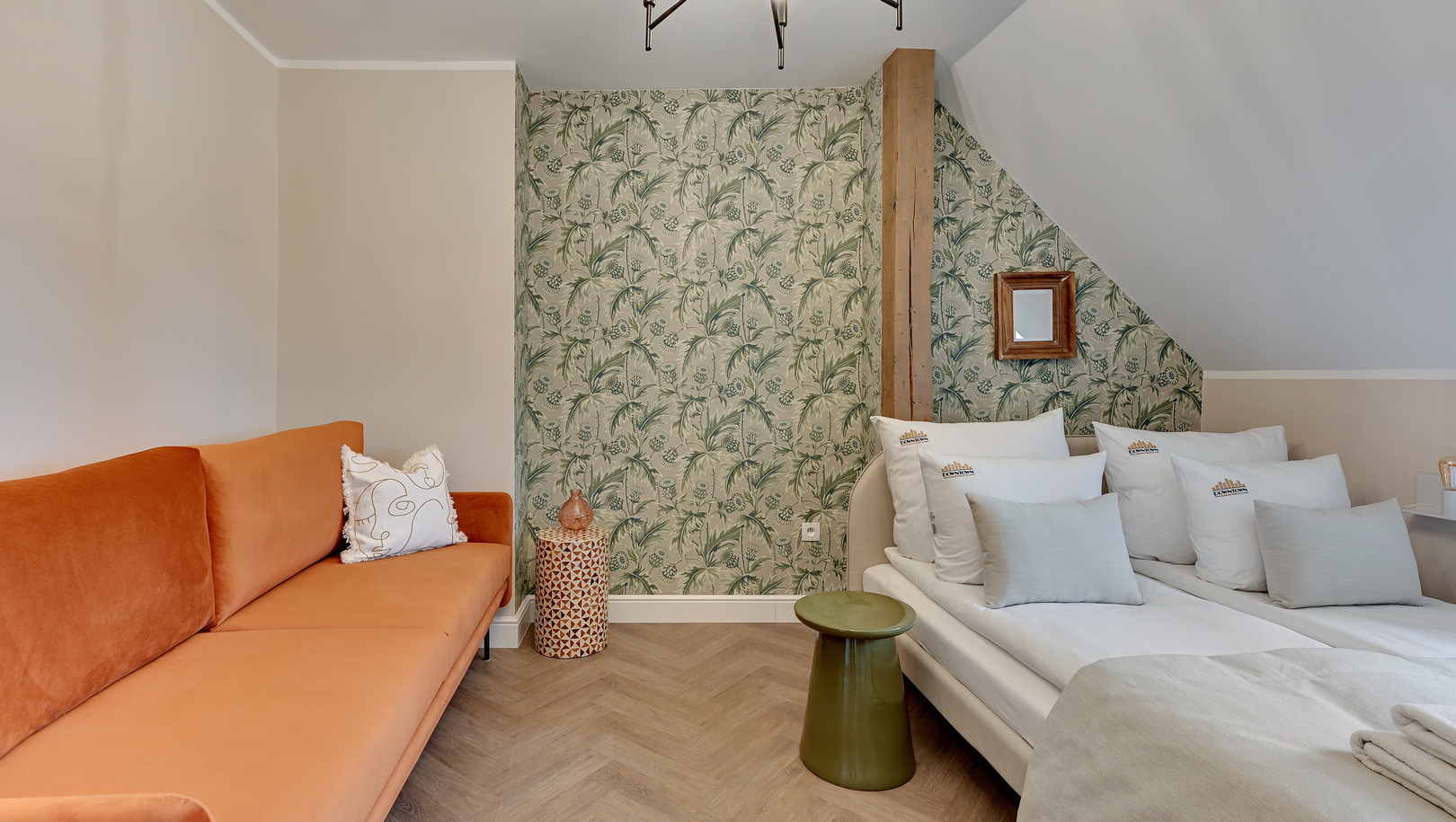Przytulny pokój gościnny z łóżkami połączonymi w jedno, miękkimi białymi poduszkami, pomarańczową sofą, botaniczną tapetą, drewnianą belką i zielonym stolikiem.