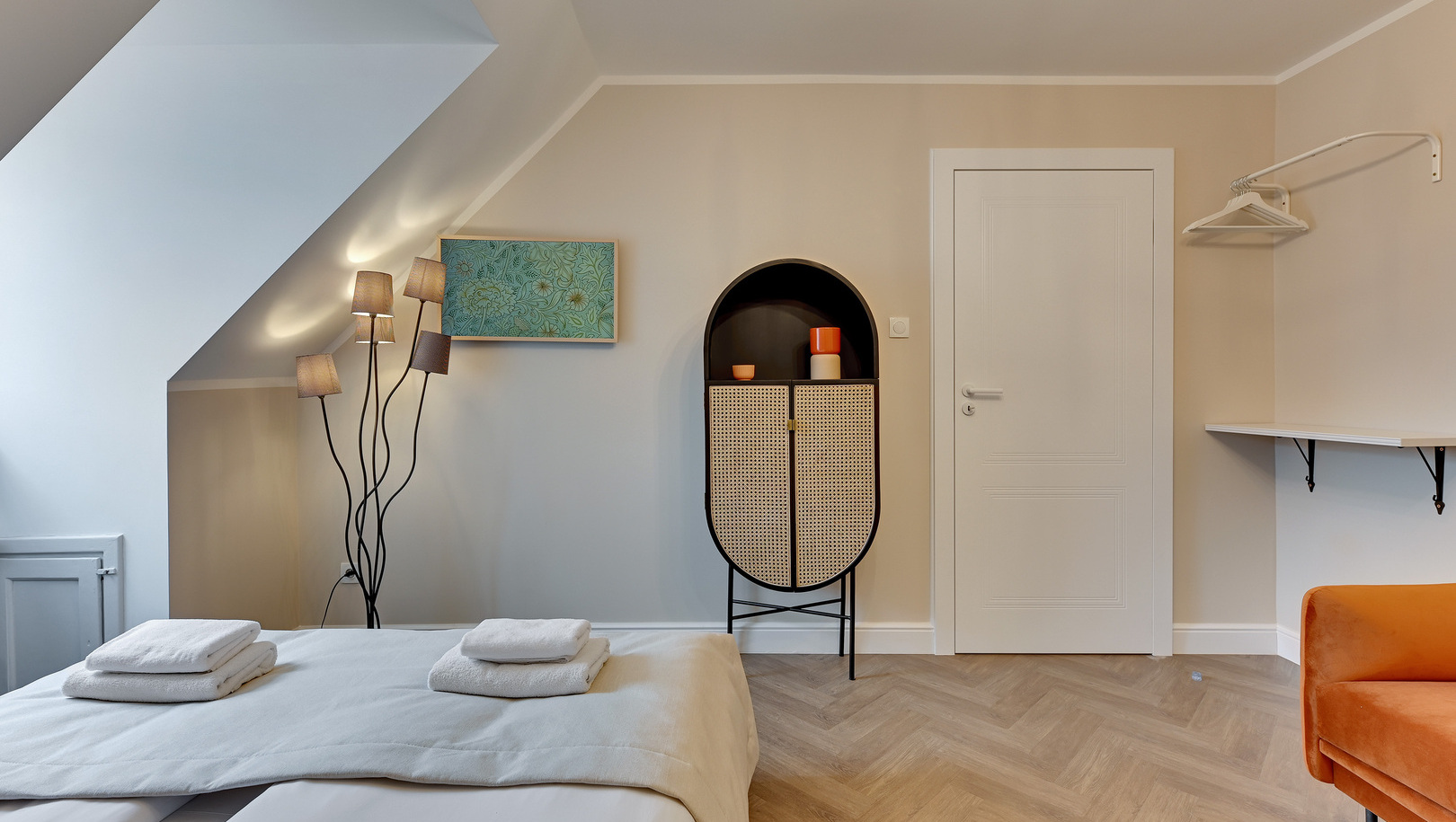 Poddasze z łóżkiem małżeńskim, starannie złożonymi ręcznikami, artystyczną lampą stojącą, rattanową szafką i pomarańczowym fotelem
