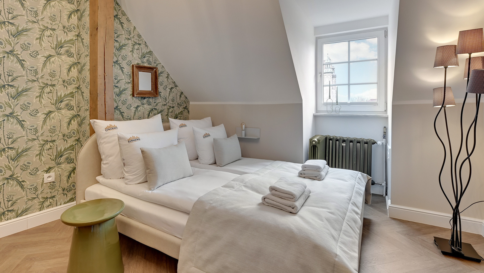 Poddasze: pokój dwuosobowy ze skosami, kwiatową tapetą, odsłoniętą belką, białą pościelą, ręcznikami i widokiem na wieżę.