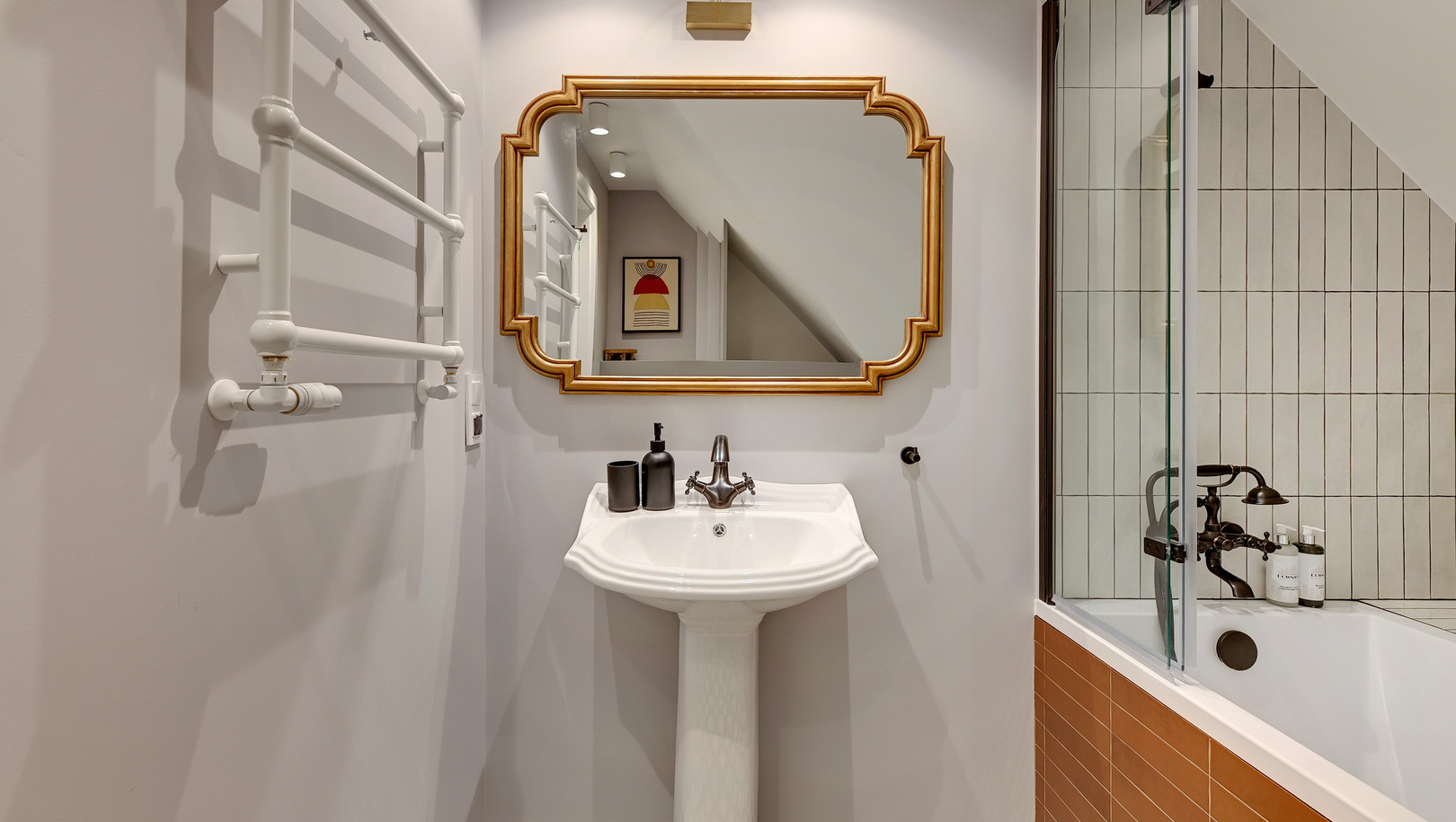 Butikowa łazienka z białą umywalką na nodze, złotym lustrem, podgrzewanym wieszakiem na ręczniki, kaflową wanną i kosmetykami