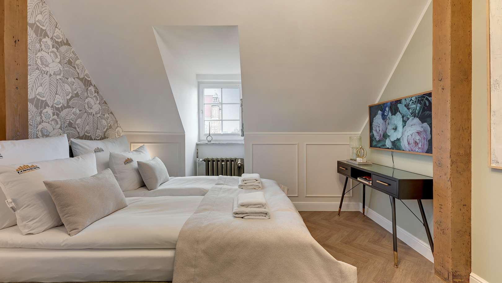 Przytulny pokój na poddaszu z połączonymi łóżkami, jasną pościelą, złożonymi ręcznikami, odsłoniętymi belkami i widokiem na miasto