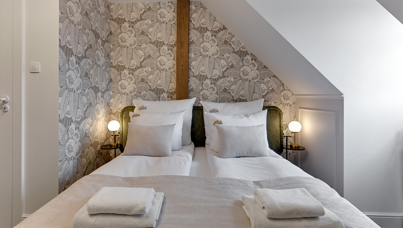 Przytulne łóżko na poddaszu z kwiatową tapetą, skośnym sufitem, zielonym wezgłowiem, lampkami nocnymi i złożonymi ręcznikami