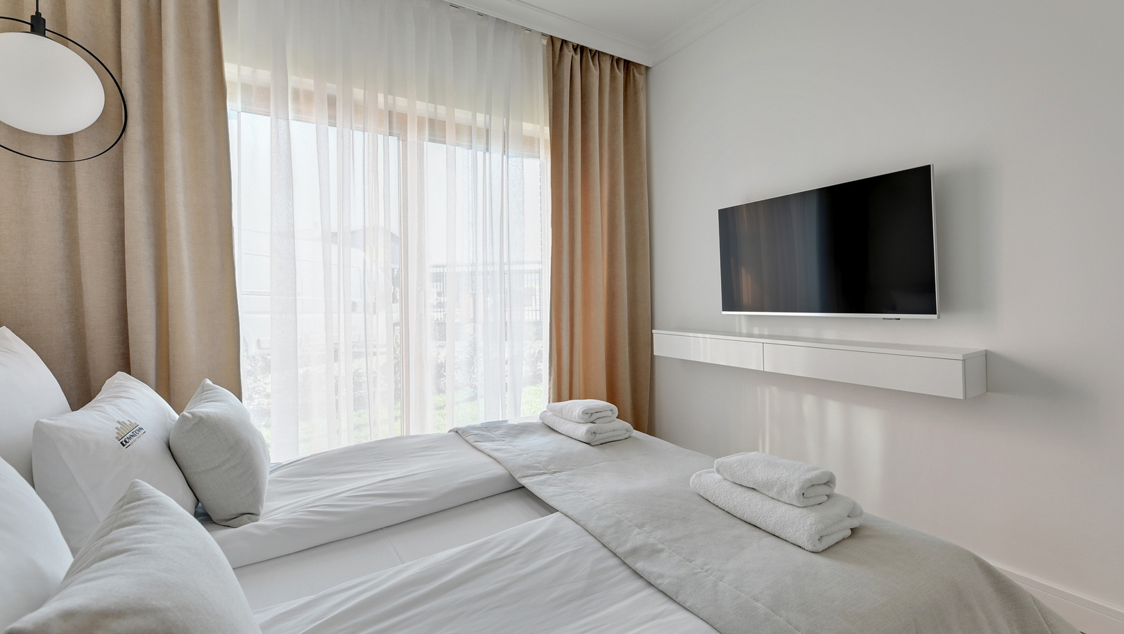 Dwa łóżka z białą pościelą, złożone ręczniki, telewizor na ścianie i duże okno z prześwitującą firaną i beżowymi zasłonami