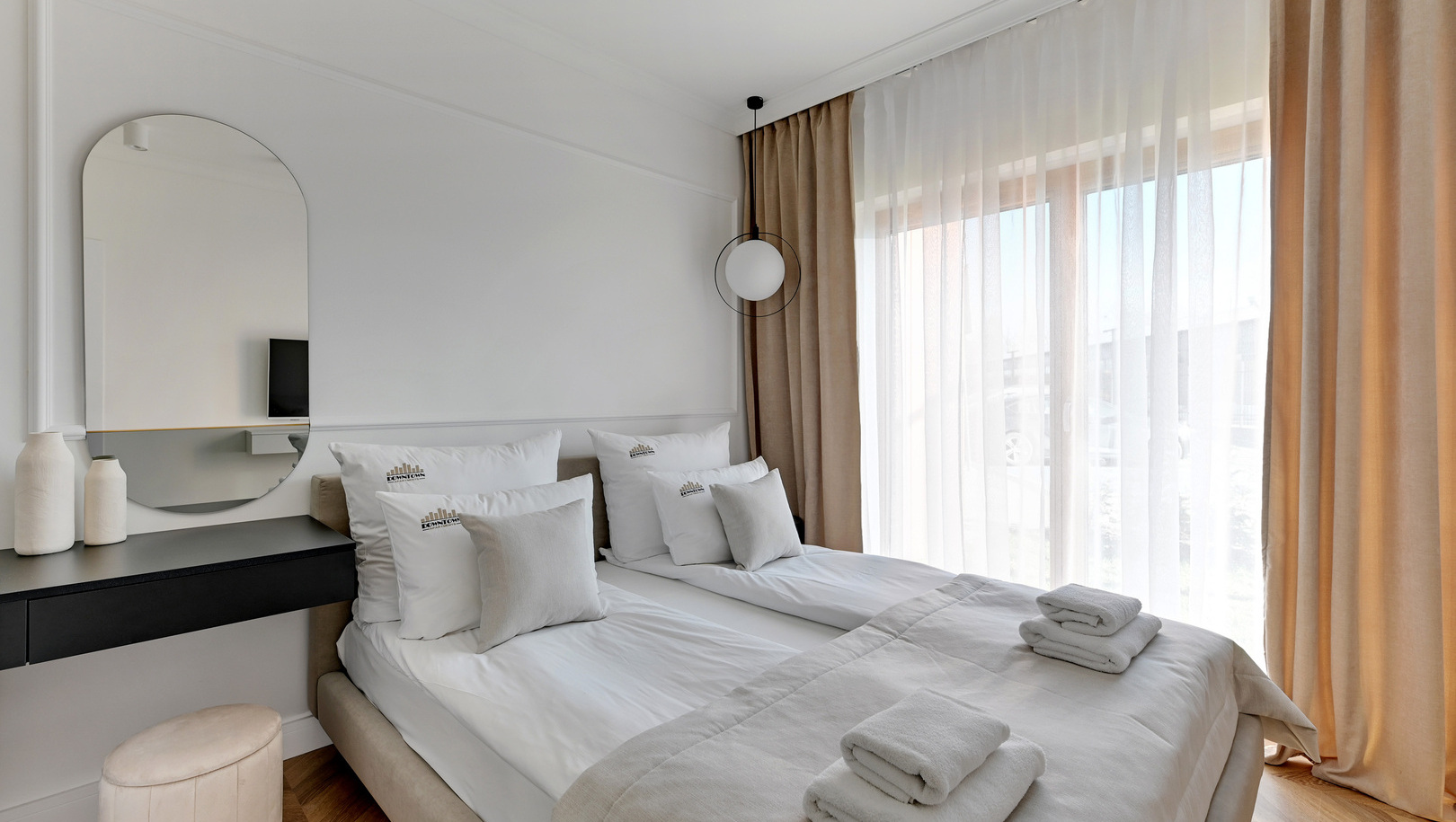 Jasny, nowoczesny pokój hotelowy z połączonymi materacami, neutralną pościelą, złożonymi ręcznikami, okrągłym lustrem i zasłonami