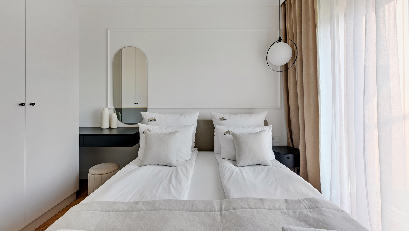 Jasny nowoczesny pokój z dwoma pojedynczymi łóżkami w białej pościeli, lampą wiszącą, toaletką, beżowymi zasłonami i ręcznikami