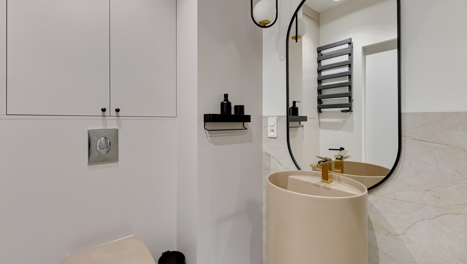 Nowoczesna łazienka dla gości z beżową wolnostojącą umywalką, złotym kranem, owalnym lustrem i grzejnikiem na ręczniki.