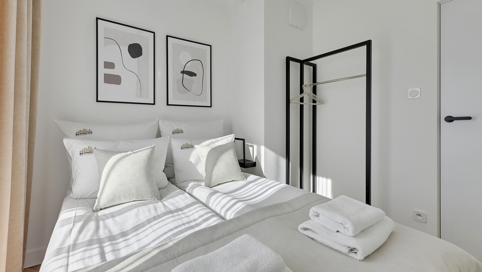 Jasny, minimalistyczny pokój z dwoma łóżkami, białą pościelą, złożonymi ręcznikami, abstrakcyjną grafiką i stelażem na ubrania