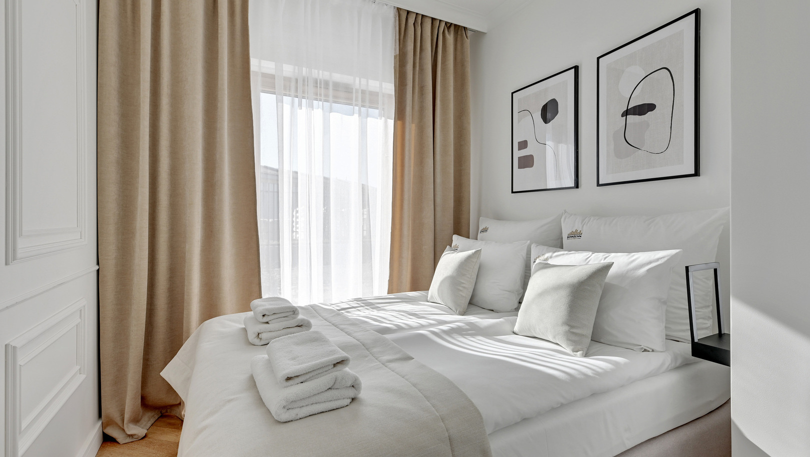 Jasny, minimalistyczny pokój z dużym łóżkiem, świeżą białą pościelą, złożonymi ręcznikami, beżowymi zasłonami do podłogi, tiulem i abstrakcyjnymi obrazami