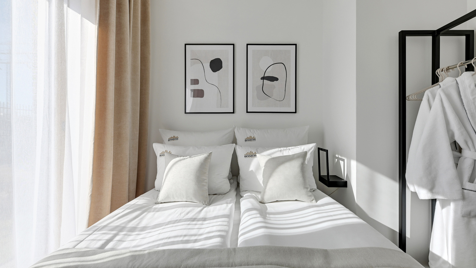 Jasne, minimalistyczne złączone łóżka, biała pościel, abstrakcyjne obrazy, przezroczysta i beżowa zasłona, miękkie szlafroki na stojaku.