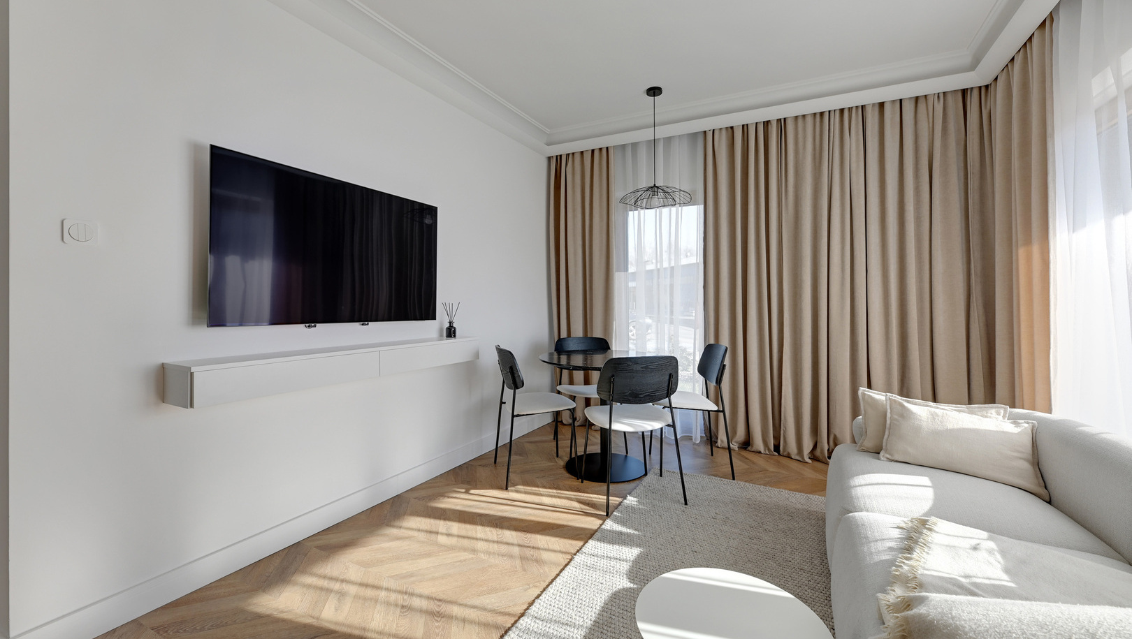 Jasny nowoczesny salon hotelowy z telewizorem na ścianie, stołem z krzesłami, sofą i beżowymi zasłonami od podłogi do sufitu.