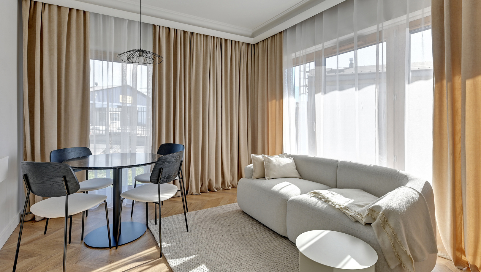 Nasłoneczniony salon z zaokrągloną kremową sofą, przytulnym kocem, okrągłym stołem na 4 krzesła, firany do podłogi i drewniana podłoga