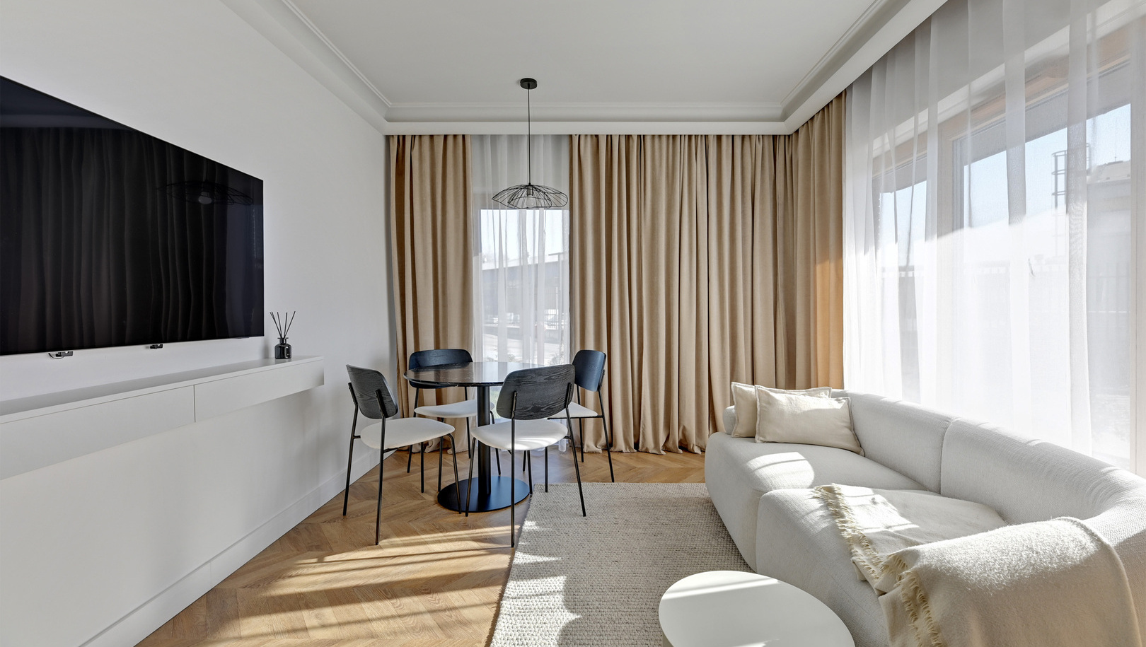 Jasny, nowoczesny salon w apartamencie z białą sofą, pledem, dywanem, stołem z czterema krzesłami, telewizorem i wysokimi oknami z woalami