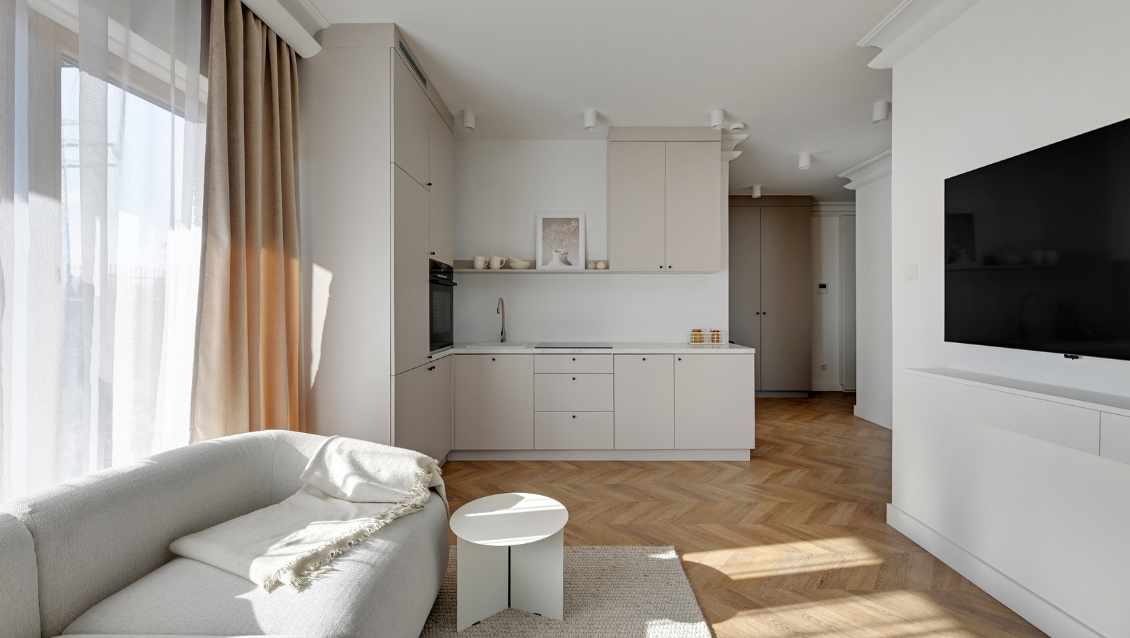Jasne studio z aneksem kuchennym, sofą, małym okrągłym stolikiem, TV, zasłonami do podłogi i parkietem w jodełkę.