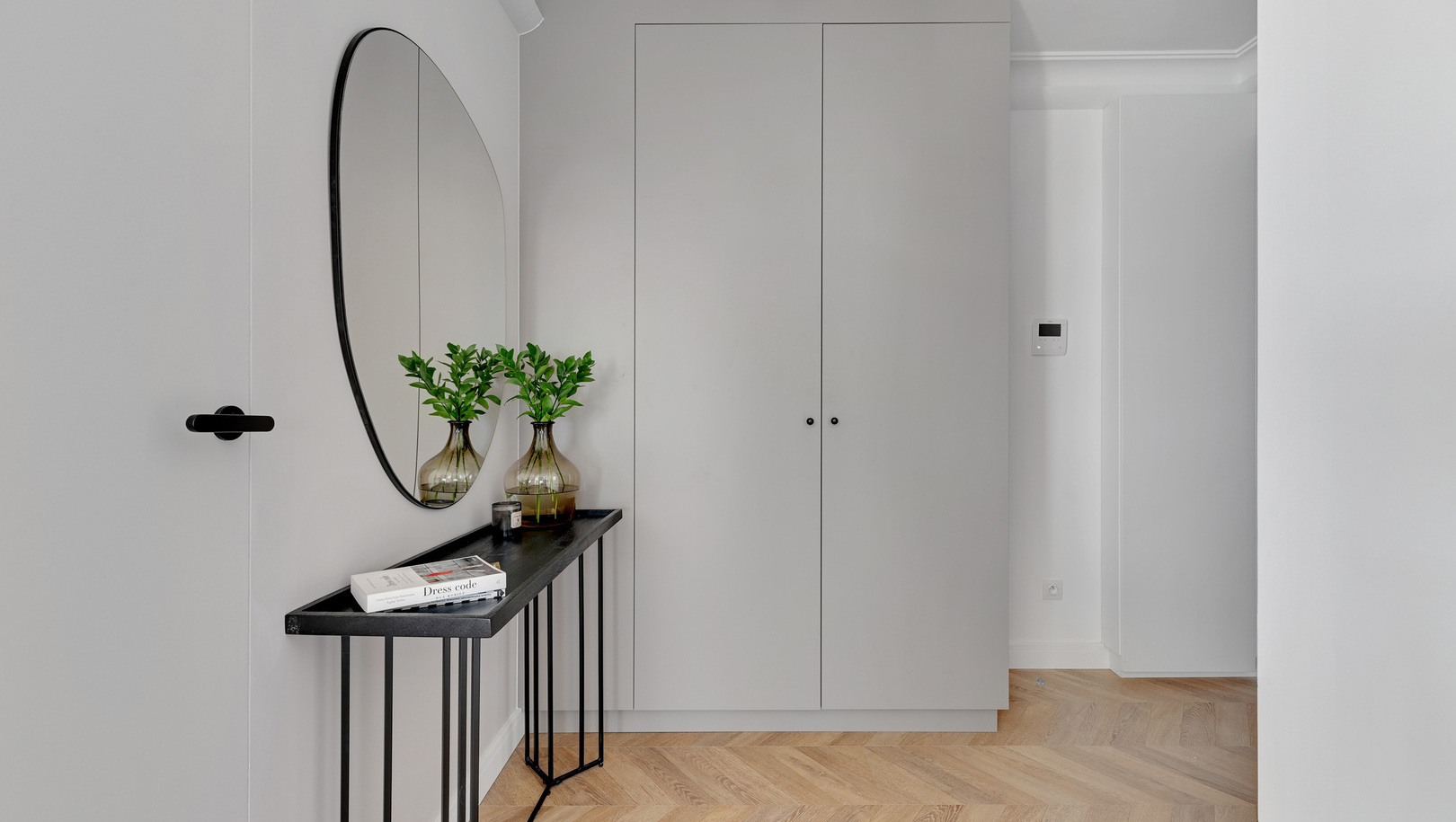 Minimalistyczne wejście do apartamentu z wbudowaną szafą, czarną konsolą, okrągłym lustrem i dębową podłogą