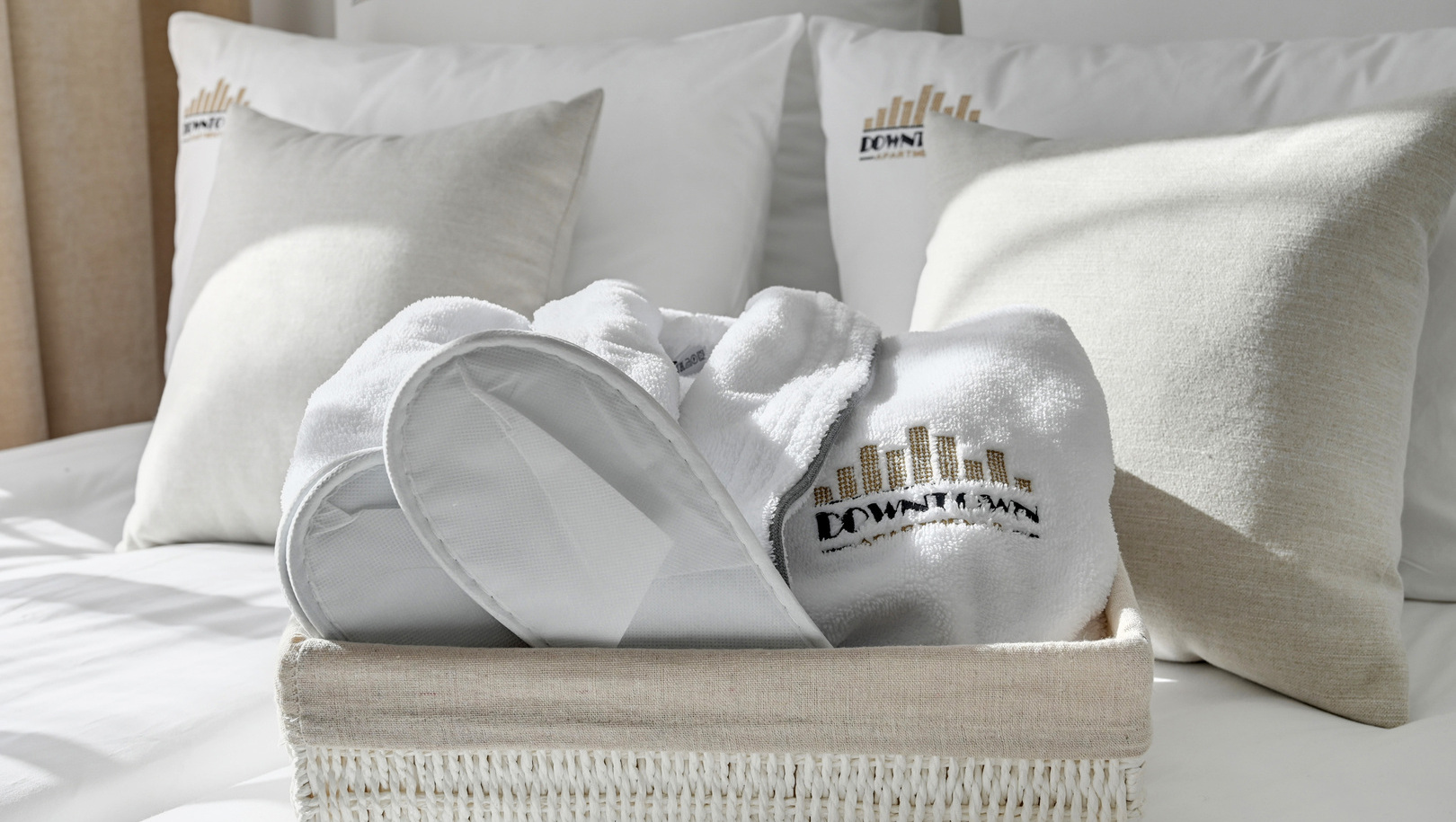 Biały puszysty szlafrok i kapcie z wyszywanym logo hotelu w plecionym koszu na białym łóżku z miękkimi poduszkami.