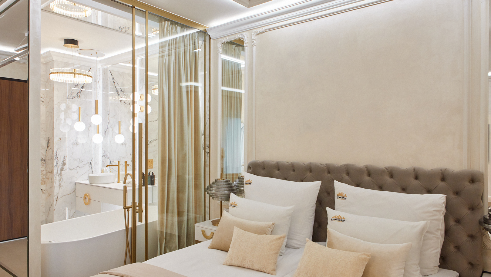 Luksusowy apartament z pikowanym zagłówkiem, miękkimi poduszkami, beżową pościelą i szklaną ścianką łazienki z wolnostojącą wanną i złotymi armaturami