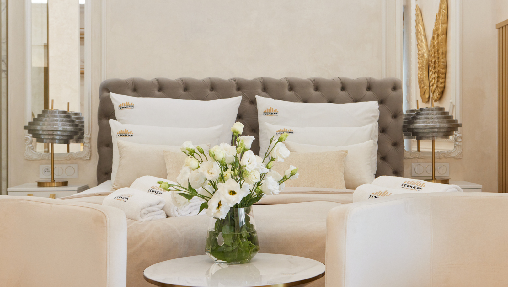 Elegancki pokój w neutralnych odcieniach z pikowanym zagłówkiem, poduchami, marmurowym stolikiem z białymi kwiatami, fotelami i hotelowymi szlafrokami.