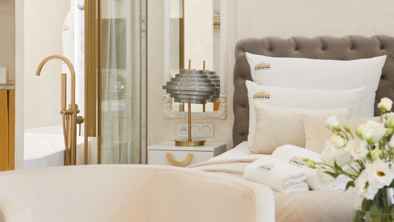 Łóżko z pikowanym zagłówkiem, miękka biała pościel, lampa przy stoliku, złota bateria wolnostojąca, świeże kwiaty i zwinięte ręczniki