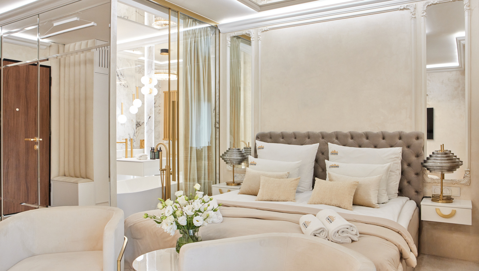 Luksusowy apartament w neutralnych tonach z pikowanym łóżkiem king, aksamitnymi fotelami, świeżymi kwiatami, zwiniętymi ręcznikami i przeszkloną marmurową łazienką